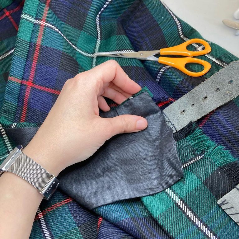 kilt expert alteration glasgow A hand holding a black fabric patch on a tartan fabric, with scissors nearby.